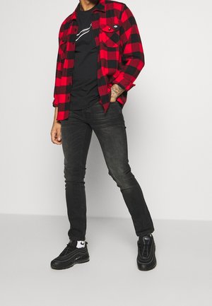 Mężczyzna w czerwono-czarnej koszuli w kratę na czarnym t-shircie z grafiką, czarnych obcisłych jeansach, czarnych trampkach i białych skarpetkach, stojący na białym tle.