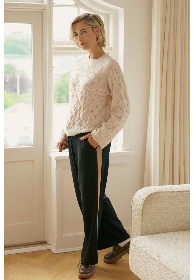 Kremowy sweter z dzianiny o fakturowanym wzorze w romby, zestawiony z czarnymi spodniami o szerokich nogawkach z bocznym beżowym paskiem. Na podłodze stoją brązowe buty.