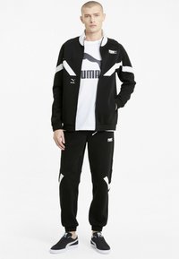 Fekete sportos melegítő, kontrasztos fehér csíkokkal, cipzáros dzsekivel és tapered nadrággal, sima anyagból készült. Fehér logó látható.