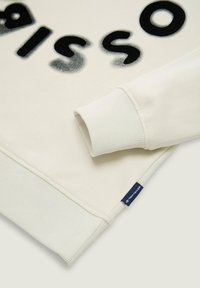 Sudadera blanca con textura acanalada, que presenta un gran texto negro. Los puños y el dobladillo también son acanalados. Una etiqueta de logotipo azul es visible en el lateral.