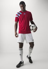Rotes Sportshirt mit blauen Akzenten, weiße Shorts, schwarze Fußballschuhe und knielange weiße Socken mit schwarzen Streifen. Hält einen Fußball.