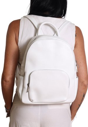 Vrouw in een mouwloze witte top en broek, met een witte leren rugzak met een voorzak met rits en zijzakken.