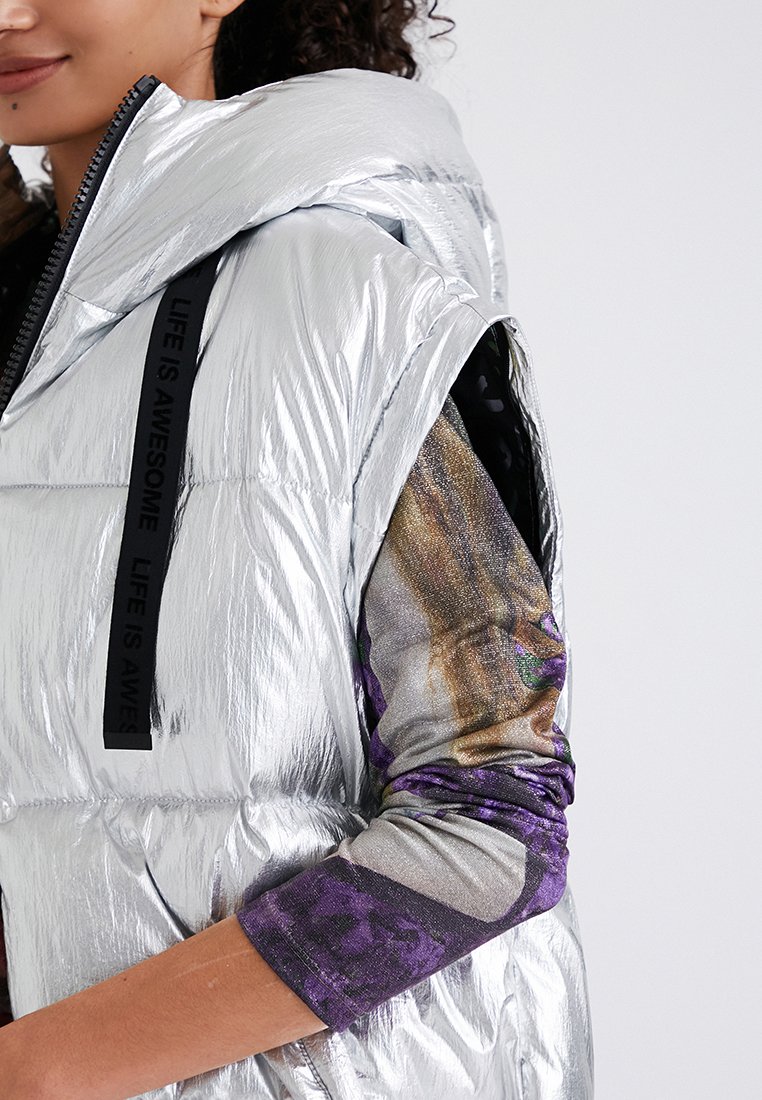 Kobieta w błyszczącej srebrnej kamizelce bez rękawów, nosi długi rękaw w wielobarwnym topie w odcieniach fioletu i złota.