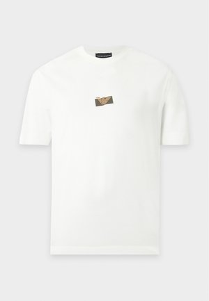 Άντρας που φοράει λευκό κοντομάνικο t-shirt με μικρό λογότυπο, μπλε τζιν και λευκά αθλητικά, στέκεται μπροστά σε έναν απλό φωτεινό φόντο.
