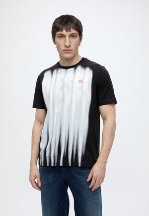 Homme aux cheveux courts et foncés porte un t-shirt noir avec des rayures blanches verticales et un grand texte flou « DIESEL », associé à un jean bleu.
