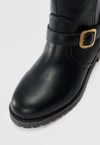 Bota de cuero negra hasta el tobillo con punta redonda, acento de hebilla dorada, detalles de costura y suela de goma texturizada.