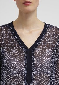 Czarna i biała bluzka wzorzysta z dekoltem w serek, przezroczystym materiałem i jednolitym czarnym wykończeniem wzdłuż dekoltu i listwy guzikowej.