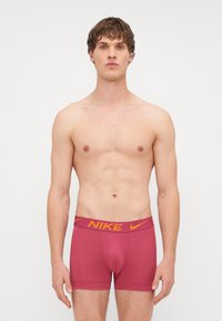 Crvene Nike bokserice s žutim logoom branda na pojasu. Materijal je gladak, s uskim fitom i minimalnim šavovima.