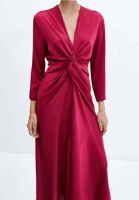 Donna che indossa un vestito a maniche lunghe di un rosso intenso con un design a nodo torciglione in vita e scollatura a V, su uno sfondo semplice.