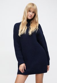 Tumši zila adīta džemperkleita ar augstu apkakli, ribotām detaļām piedurknēs un apakšmalā, un vaļēju piegriezumu, kas izceļ auduma tekstūru.