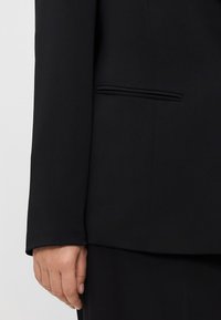 Črn blazer z gladko teksturo, opremljen z enim žepom na robu. Rokavi so dolgi, celoten dizajn pa je prilagojen in strukturiran.