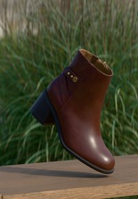 Stivale alla caviglia in pelle marrone con punta leggermente affusolata, zip laterale e cinturino decorativo con dettagli dorati. Tacchetto largo e resistente.