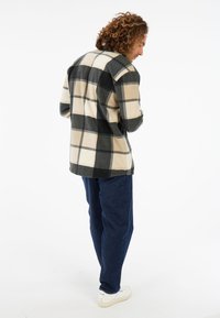 Man met krullend haar, gekleed in een zwart en beige groot geruit jasje, donkere jeans en witte sneakers, staand en naar beneden kijkend.