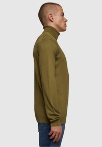 Olivgrön polotröja i mjukt, strukturerat tyg; åtsittande design med ribbad mudd och nederkant; kombinerad med blå jeans; sidovy.