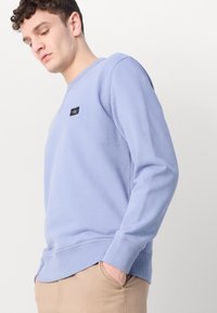 Lysblå sweatshirt med ribbede manchetter og kant, der har en lille sort logo-patch på venstre bryst. Blødt stof og afslappet pasform.