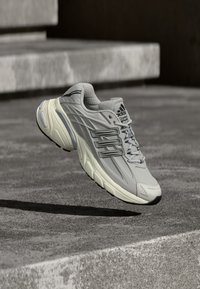Grey running shoe with a textured mesh upper, rubber sole, and three lateral stripes. Features a padded collar and reinforced toe cap.
