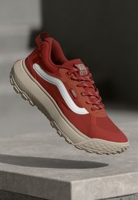 Roter Vans MTE Sneaker mit beigefarbener Sohle und weißem Seitenstreifen, in der Luft über einer Betonfläche schwebend dargestellt.