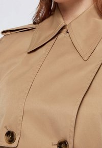 Beżowy płaszcz trencz z dużym kołnierzem, dwurzędowym zapięciem na guziki i gładką fakturą. Widoczne przeszycia wzdłuż szwów.