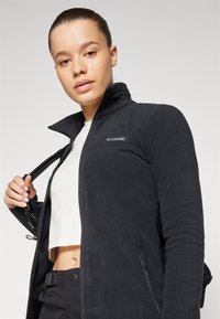 Must värviline fleece-jakk, millel on silmadega krae, külgmised lukuga taskud ja väike logo rinnal. Sile tekstuur ja sobiv disain.