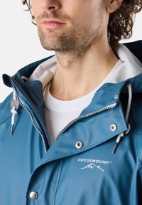 Blå vattentät jacka med en ljusgrå huva, silverfärgade tryckknappar och justerbara dragsnören. Har en logotyp på bröstet och en slät yta.