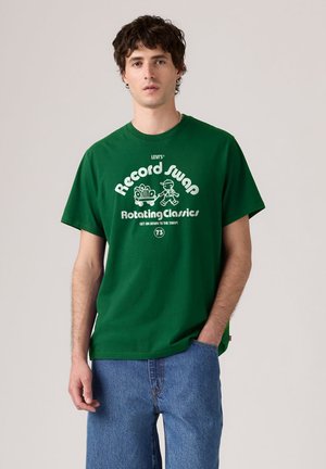 Homme portant un T-shirt vert Levi's avec le texte et le graphisme "Record Swap Rotating Classics", associé à un jean bleu, debout devant un fond uni.