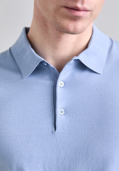 Svetlomodrá polo košeľa vyrobená z mäkkej tkaniny, s klasickým golierom a trojzubkovou légaou. Hladká textúra bez viditeľných vzorov.