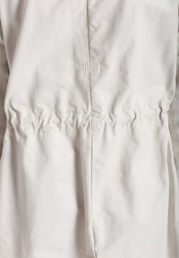 Giacca beige chiaro realizzata in tessuto morbido, con vita arricciata e ricamo discreto "Khujo" vicino alla parte superiore. Texture liscia.