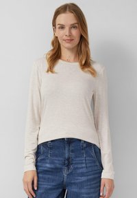 Blondynka z niebieskimi oczami ma na sobie jasnobeżową bluzkę z długim rękawem i niebieskie dżinsy z wysokim stanem, stoi na tle jednolitego jasnego tła.