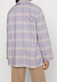 Femme portant un blazer ample à carreaux dans des couleurs pastel douces et un pantalon en velours côtelé beige, vue de dos.