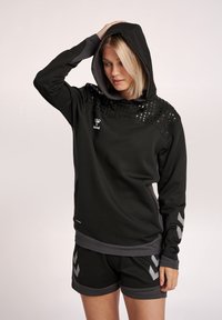 Felpa nera con una texture a pois nella parte superiore, dettagli grigi e un logo frontale. Abbinata a pantaloni corti neri con motivi a zig-zag grigi.