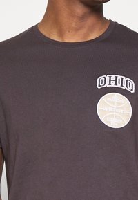 Homme portant un T-shirt gris foncé avec le texte « OHIO » et un écusson graphique de basket-ball indiquant « Ohio Brooklyn University Champ 2014 » sur la poitrine.