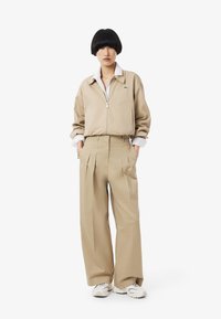Giacca beige con zip e polsini a coste, abbinata a pantaloni beige a vita alta e plissettati. Collo bianco visibile, accentuato da scarpe da ginnastica.