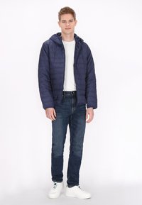 Marine blauwe pufferjack met een capuchon, gevoerd met een quilted textuur, gedragen over een wit t-shirt, gecombineerd met blauwe jeans en witte sneakers.