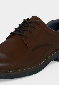 Chaussure habillée en cuir marron avec des lacets noirs, des coutures visibles et une semelle en caoutchouc noire sur un fond clair et uni.