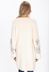 Crème-kleurige lange cardigan, gebreide textuur, met bloemenborduursels op de mouwen, ontspannen pasvorm, verlaagde schouders en lengte tot halverwege de dijen.