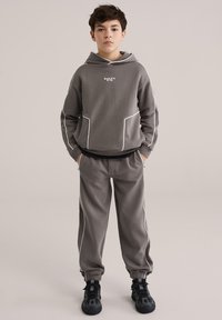 Hanorac gri cu glugă și detalii albe, prevăzut cu buzunare frontale; asortat cu pantaloni cu manșete în aceeași nuanță. Purtați cu pantofi sport negri.