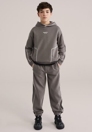 Felpa grigia con cappuccio e piping bianco con tasche frontali; abbinata a pantaloni coordinati con polsini. Indossata con scarpe da ginnastica nere.