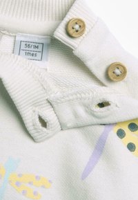 Polo shirt per neonati color crema con due bottoni rotondi in legno, colletto a costine, dettaglio a piccoli fori e motivi di insetti colorati sul tessuto.