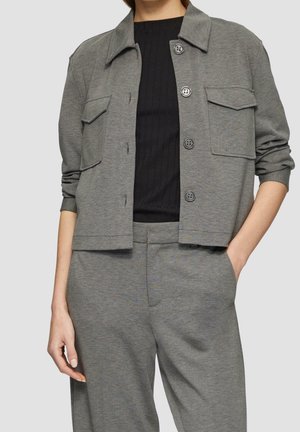 Femme portant une veste grise boutonnée avec des poches à rabat, un haut noir côtelé et un pantalon gris assorti, une main dans la poche.