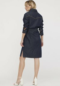 Robe en denim bleu foncé avec des manches longues, taille ceinturée et fentes latérales. Elle présente des détails cousus sur l'empiècement arrière et des poignets boutonnés.