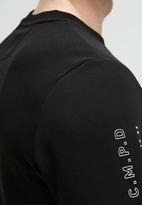Black long-sleeve shirt featuring a textured shoulder, with white branding printed vertically along the arm. Made of a smooth, stretch fabric.