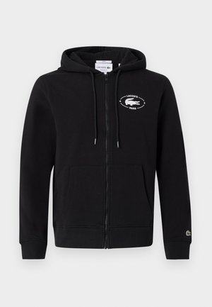 Twee mensen dragen zwarte hoodies met ritssluiting en witte logo's, gecombineerd met lichtblauwe wijde jeans, staand tegen een effen witte muur.
