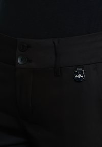 Czarne spodnie z dwoma czarnymi guzikami, gładką fakturą i metalicznym akcentem w okrągłym kształcie blisko pasa.