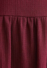 Burgundowa sukienka o teksturowanej powierzchni z marszczonym dołem, z pionowymi paskami i marszczonym gorsami. Miękki materiał o delikatnym połysku.