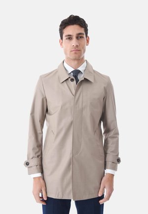 Uomo che indossa un trench beige, camicia bianca, cravatta blu navy a pois e pantaloni scuri, in piedi davanti a uno sfondo bianco uniforme.