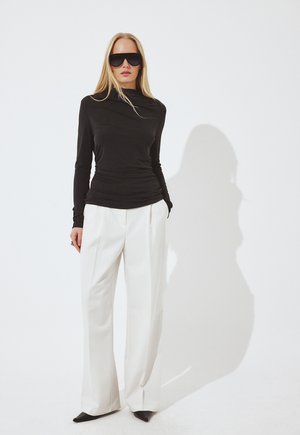 Naine, kes kannab suuri musti päikeseprille, musta pika varrukaga pluusi, valgeid laiade säärtega pükse ja musti terava ninaga jalanõusid valge tausta ees.