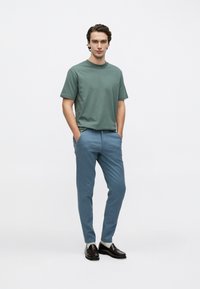 Πράσινο βαμβακερό t-shirt και μπλε chinos, με άνετη εφαρμογή. Μαύρα παπούτσια με υφή, χέρια στις τσέπες, ουδέτερο φόντο.