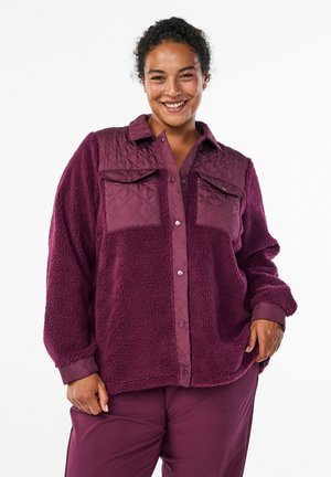 Lila gesteppte Hemdjacke mit flauschiger Textur, ausgestattet mit zwei aufgesetzten Taschen, Knopfschluss und gerippten Bündchen. Ideal zum Layering.