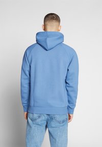 Sudadera azul con capucha ajustable, puños y dobladillo acanalados, y mangas raglán. Tejido suave con una textura lisa, combinada con jeans azul claro.