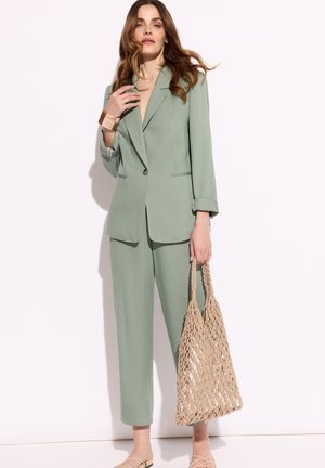 Kobieta w zielonym garniturze w kolorze szałwii, trzymająca dużą beżową plecioną torbę, nosząca bransoletkę i sandały z odkrytymi palcami na białym tle.